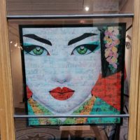 Photographie présentant le tableau "ETSUMI" (une geisha sur fond de bande dessinée) encadré. Photo prise derrière une vitrine.