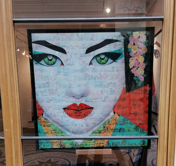 Photographie présentant le tableau "ETSUMI" (une geisha sur fond de bande dessinée) encadré. Photo prise derrière une vitrine.